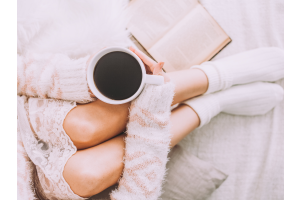 lady holding a cup of black coffee, wearing cream cardigan, cream nightshirt and cream socks, with a book.