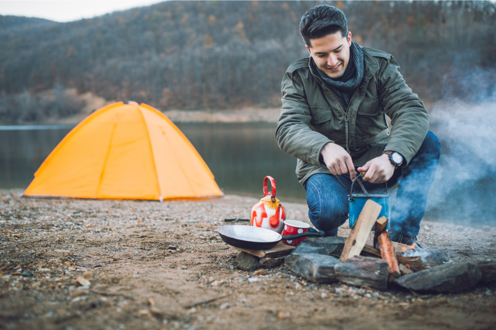 man cooking over open fire in front of an orange tent by a lake