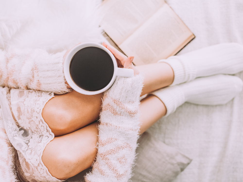 lady holding a cup of black coffee, wearing cream cardigan, cream nightshirt and cream socks, with a book.