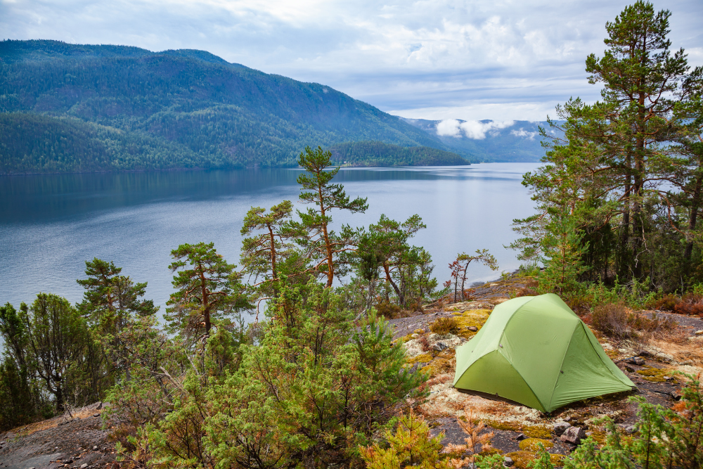 green tent on the side of a lake surrounded by trees with hills in the background