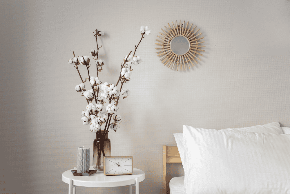 a bed with white cotton bedding, next to a white nightstand with a vase containing cotton plants 